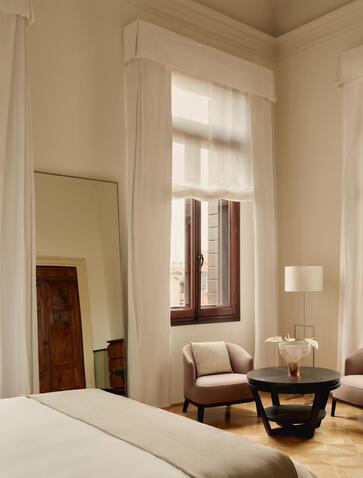 Bedroom with frescoed ceiling at Aman Venice, featuring twin reading chairs by tall windows and neutral décor.
