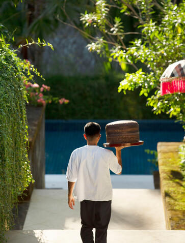 Private butler at Aman Villas at Nusa Dua, Indonesia, carrying refreshments through a shaded courtyard.