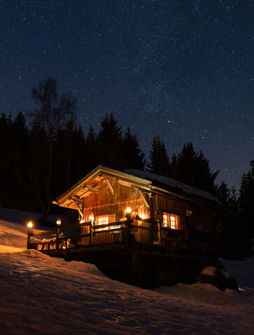 Wooden chalet glowing warmly at dusk, surrounded by snow and evergreen forest at Aman Le Mélézin, France.
