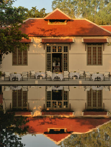 Amantaka's poolside dining table set for dinner beneath an orange-roofed colonial building reflected in still water.