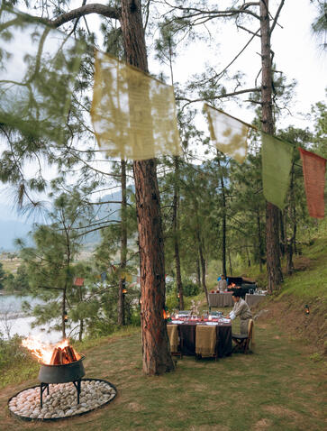 Riverside barbecue lunch at Amankora's Punakha Lodge, with guests gathered around a fire pit on the grass beneath tall trees.