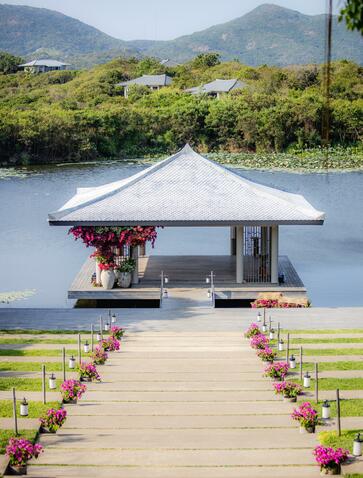 Wooden deck with pink floral arrangements leading to a white pavilion on the water at Amanoi, Vietnam, set against forested mountains.