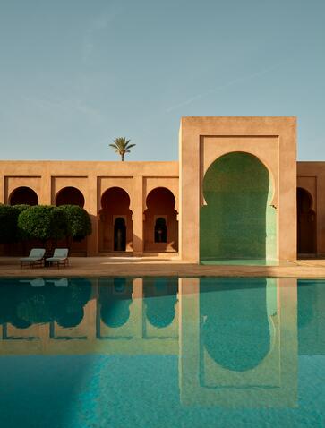 Amanjena's main pool reflects terracotta arches and a single palm tree under clear Moroccan skies.