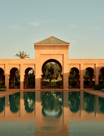 Amanjena's main pool reflects terracotta arches and a single palm tree under clear Moroccan skies.