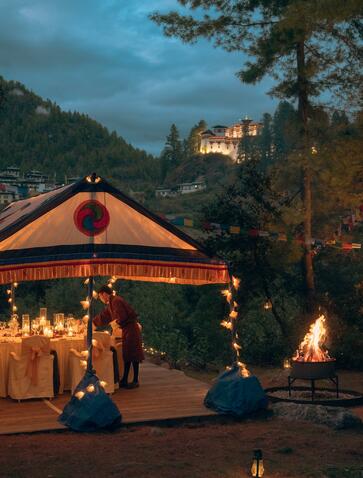 Barbecue dinner beneath Paro Taktsang fortress at Amankora, Bhutan, with glowing tent and fire at dusk.