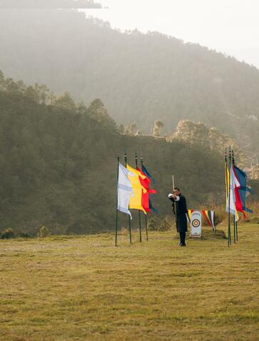 Archer practising traditional Bhutanese archery at Amankora in Punakha, with national flags and misty mountains beyond.