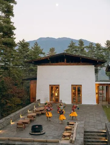 Bhutanese monks in saffron robes gather in the courtyard of Amankora's Thimphu lodge during a cultural programme, with mountains rising beyond.