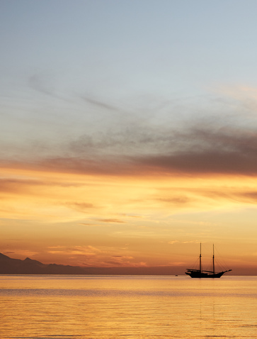Sunset over calm waters at Amandira, with a sailing vessel silhouetted against golden and orange skies.