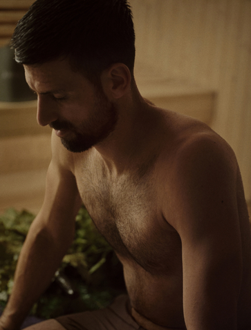 A man with dark hair sits shirtless in warm, soft lighting at Amanpuri, with green foliage visible in the background.