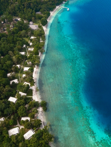 Aerial view of Amanwana's beachfront with tents nestled among tropical forest along turquoise waters, Indonesia.