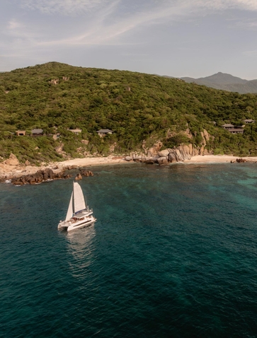 Aerial view of a white catamaran sailing off the Vietnamese coast near Amanoi, with forested hillside and sandy beach visible.