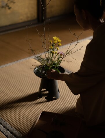 Hands arranging yellow flowers in a dark vessel at Aman Kyoto, with sunlight casting shadows across the tatami mat.