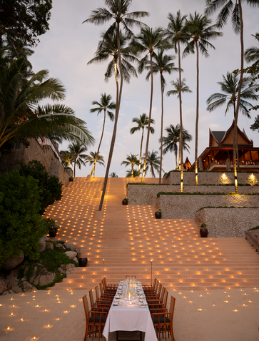 Wooden lounger on terracotta terrace overlooking Amanpuri's private beach at dusk, with palm trees and dining pavilion beyond.