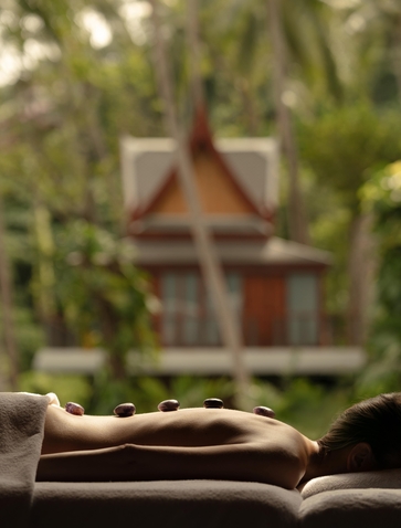 Smooth river stones arranged on a wooden surface at Amanpuri wellness spa, with tropical foliage and temple architecture blurred in the background.
