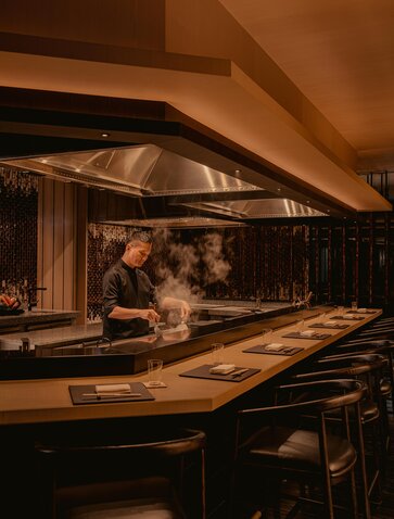 Warm-lit dining counter at Aman Nai Lert Bangkok, with chef preparing food behind glass partition.