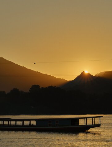 Wooden boat silhouetted on the Mekong River at sunset, mountains rising beyond at Amantaka.