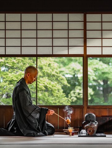 Person in dark kimono practising tea ceremony at Aman Kyoto, with garden visible through shoji screens.