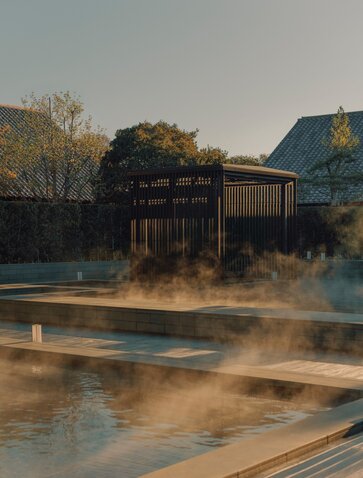 Amanemu onsen spa retreat at dawn with steam rising from thermal pools and wooden pavilion reflected in water.