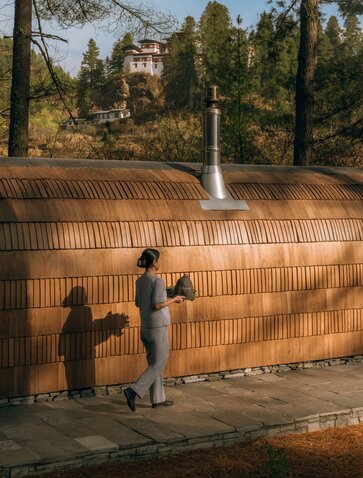Iglu sauna exterior at Amankora's Paro Lodge with two figures standing before wooden structure surrounded by cypress trees.