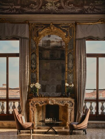 Ornate gilt-framed mirror above fireplace in Tiepolo Room at Aman Venice, with pink velvet chairs and tall windows.