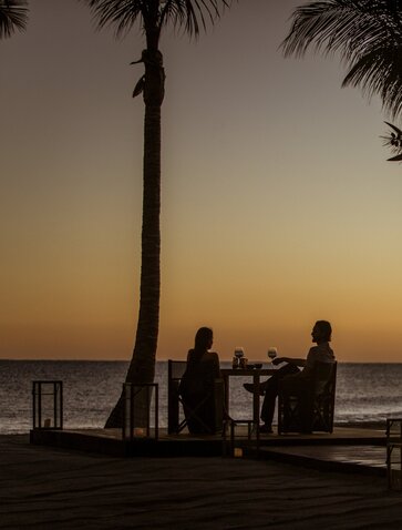 Two guests seated beneath palm trees at Amanvari, silhouetted against a golden sunset over the ocean.