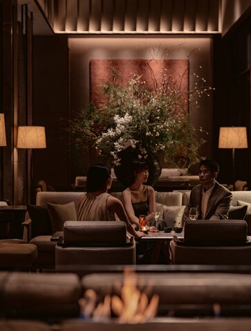 Warm-lit lounge at Aman Nai Lert Bangkok with layered seating, potted greenery, and ambient firelight.