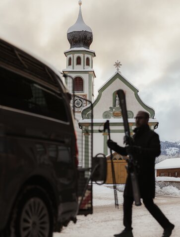 Skier boarding a shuttle at Aman Rosa Alpina, with alpine chapel visible behind in snowy Italian mountain setting.
