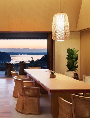 Salon et salle à manger de la villa Nagi à Amanemu, avec mobilier en bois, baie vitrée sur la mer et éclairage suspendu.
