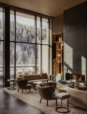 Seating area at Aman Rosa Alpina with floor-to-ceiling windows overlooking snowy Alpine forest.