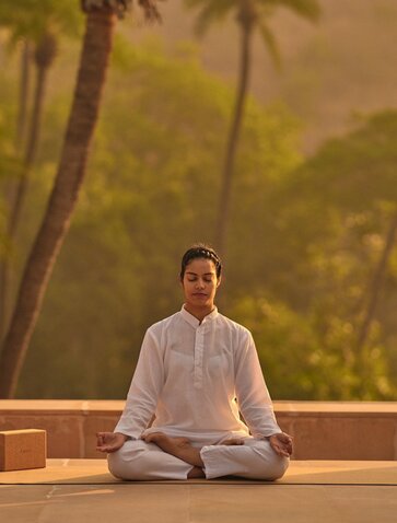 Woman meditating in lotus position at Amanbagh's spa and wellness pavilion, with palm trees visible through the window.