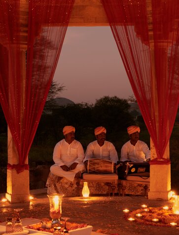 Candlelit chhatri dinner setup at Amanbagh, with three guests seated under draped red curtains surrounded by glowing candles.