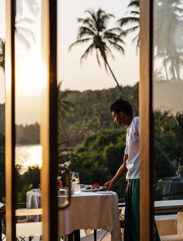 Diner seated at table on Amanwella's terrace, framed by sheer curtains, overlooking tropical palms and water.