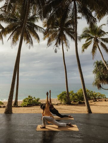 Woman practising yoga on a beachside platform at Amanwella, Sri Lanka, beneath tall palm trees.
