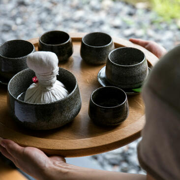 Amanemu wellness immersion: hands holding wooden tray with black ceramic bowls and white cloth, outdoor setting.