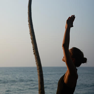 Woman practising yoga on a beach at Amangalla, reaching upward against a calm sea horizon.