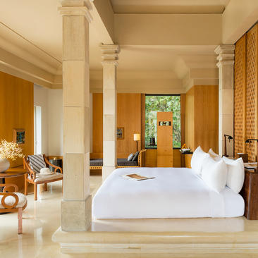 Borobudur Suite bedroom at Amanjiwo with stone columns, white bed, and view towards wooden doors opening onto Java landscape.