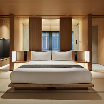 アマン京都のカエデルームの寝室。木製パネルに囲まれた広々とした空間に、低いベッドフレームの上に薄いグレーのベッドカバーが置かれている。