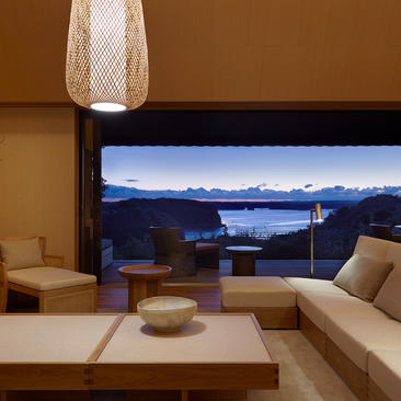 Amanemu suite with panoramic window overlooking Matsushima Bay at dusk, featuring minimalist furnishings and warm earth tones.