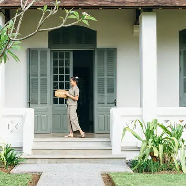 A woman stands on the stone terrace of an Amantaka suite, framed by white columns and shuttered doors, with lush greenery flanking the entrance.