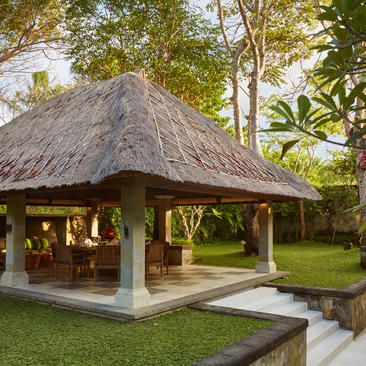 Thatched-roof dining bale at Aman Villas at Nusa Dua, surrounded by manicured gardens and tropical vegetation.