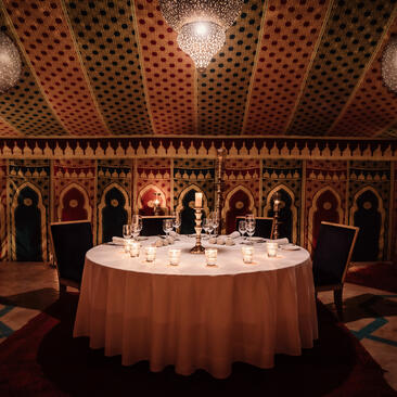 Candlelit dining room at Amanjena with round table set for service beneath ornate wooden ceiling and arched alcoves.
