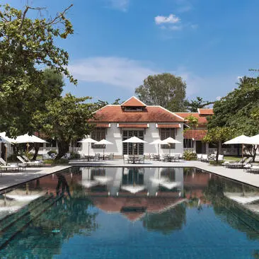 Amantaka's resort pool reflects the terracotta main building beneath clear skies, with white loungers and verdant surroundings.