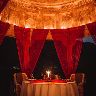 Candlelit dining table set beneath an ornate dome at Amanbagh, framed by flowing red curtains at dusk.