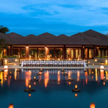 West Villa Clubhouse at Amanpulo illuminated at dusk, reflected in still water beneath twilight sky.