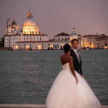 Bride and groom in wedding attire gazing at the illuminated Basilica di Santa Maria della Salute across the Venice lagoon at dusk, Aman Venice.