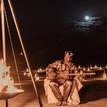 Couple seated on sand at night beneath moonlit sky at Amanjena, with lantern-lit pathways visible in background.
