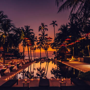 Candlelit dining setup at Amanpuri reflected in still water at dusk, palm trees silhouetted against purple sky.