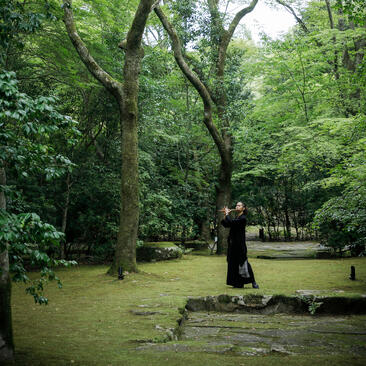 A person in dark clothing stands on a wooden bridge crossing a tranquil pond, surrounded by lush green trees at Aman Kyoto resort.