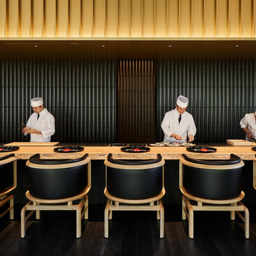 Chefs stand behind a wooden counter at Aman Kyoto's Taka An dining venue, with curved wooden chairs in the foreground.