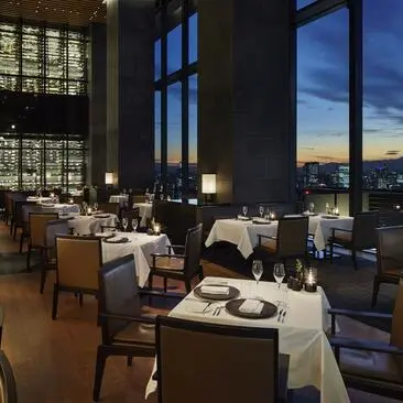 安缦东京酒店餐厅内，落地窗俯瞰城市夜景，餐桌铺设精致，夕阳映照建筑。